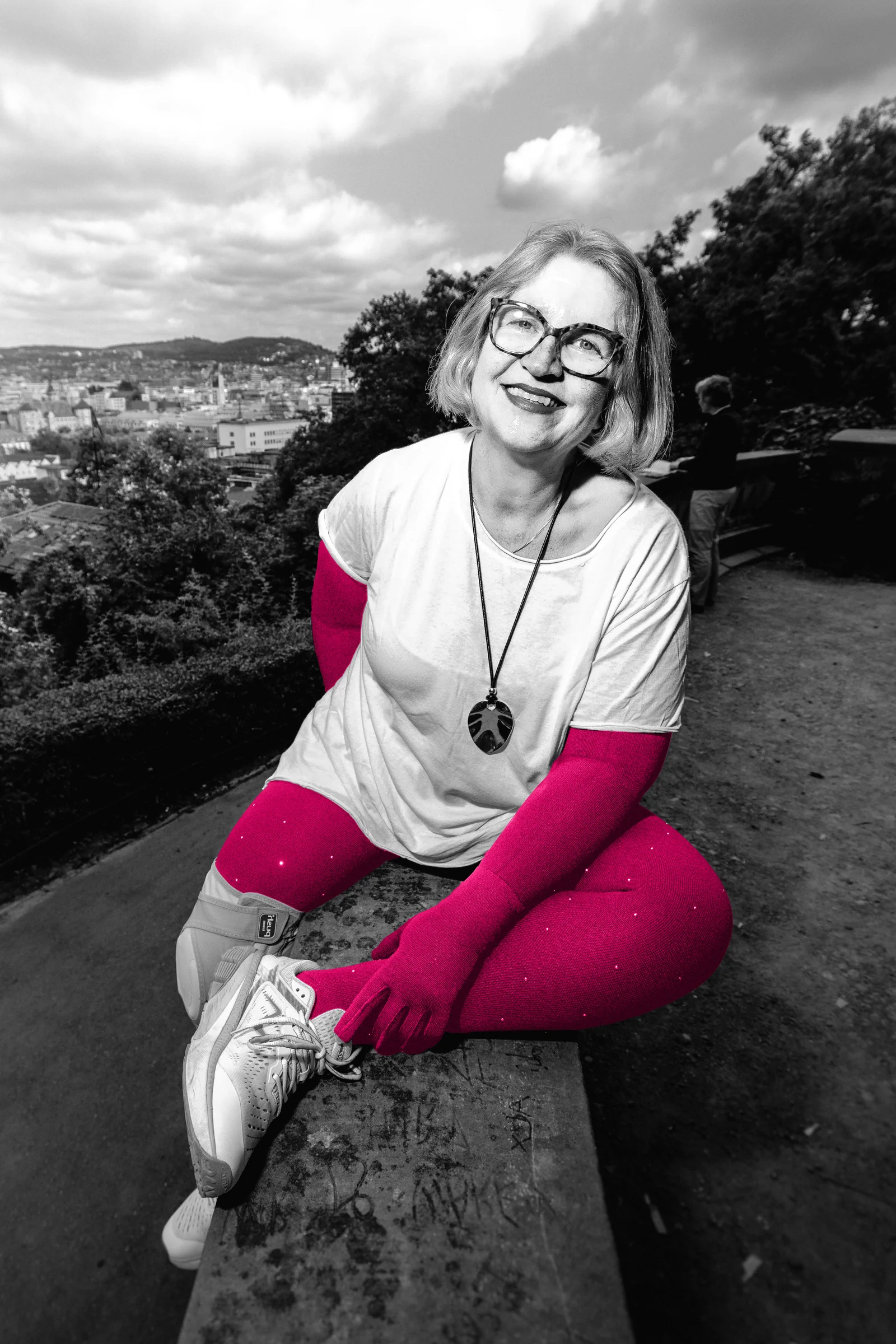 Eine Frau sitzt lachend auf einer Mauer über der Stadt, ein Bein angewinkelt.